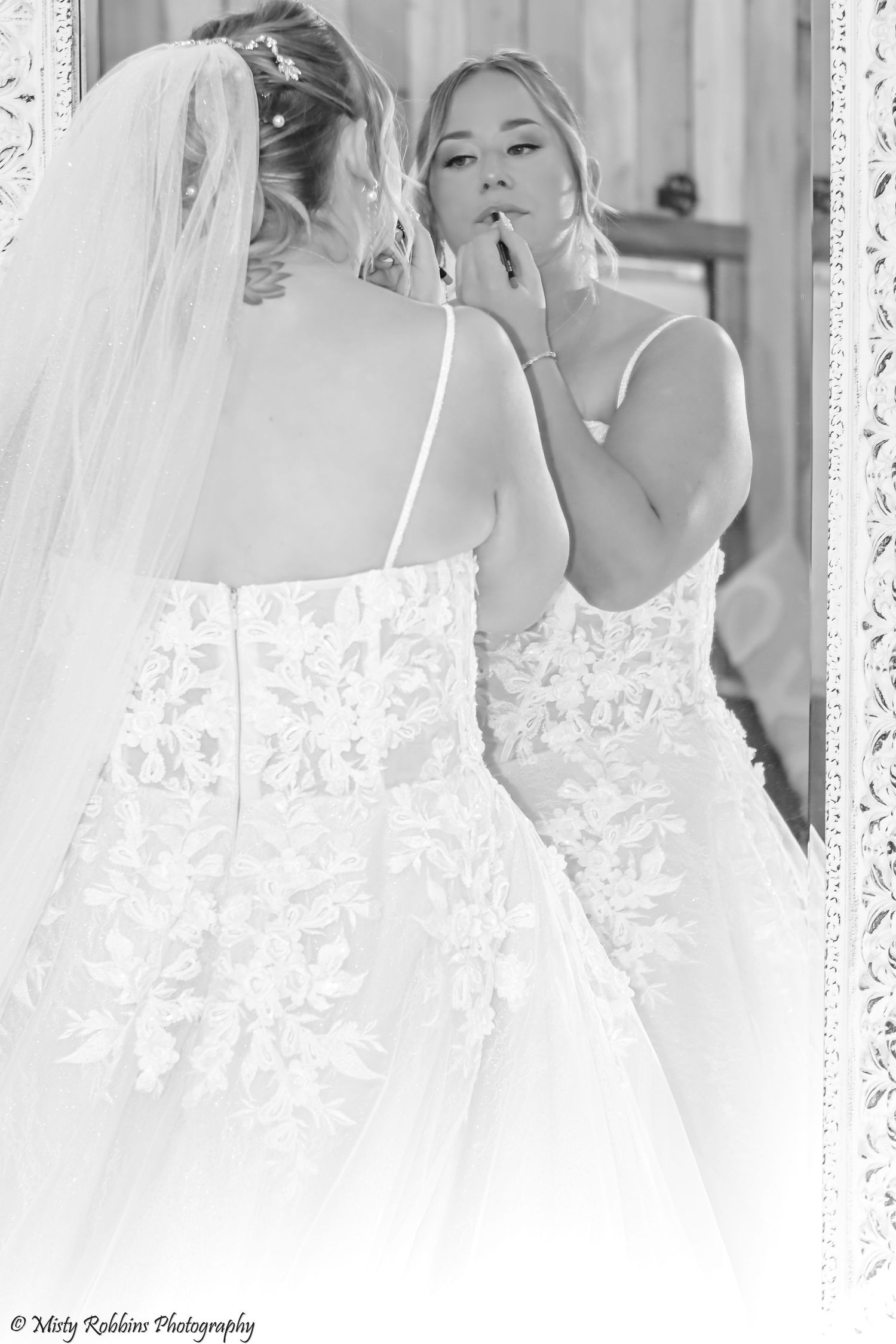 Black and white photo of a bride in a lace wedding dress looking at her reflection in an ornate mirror.