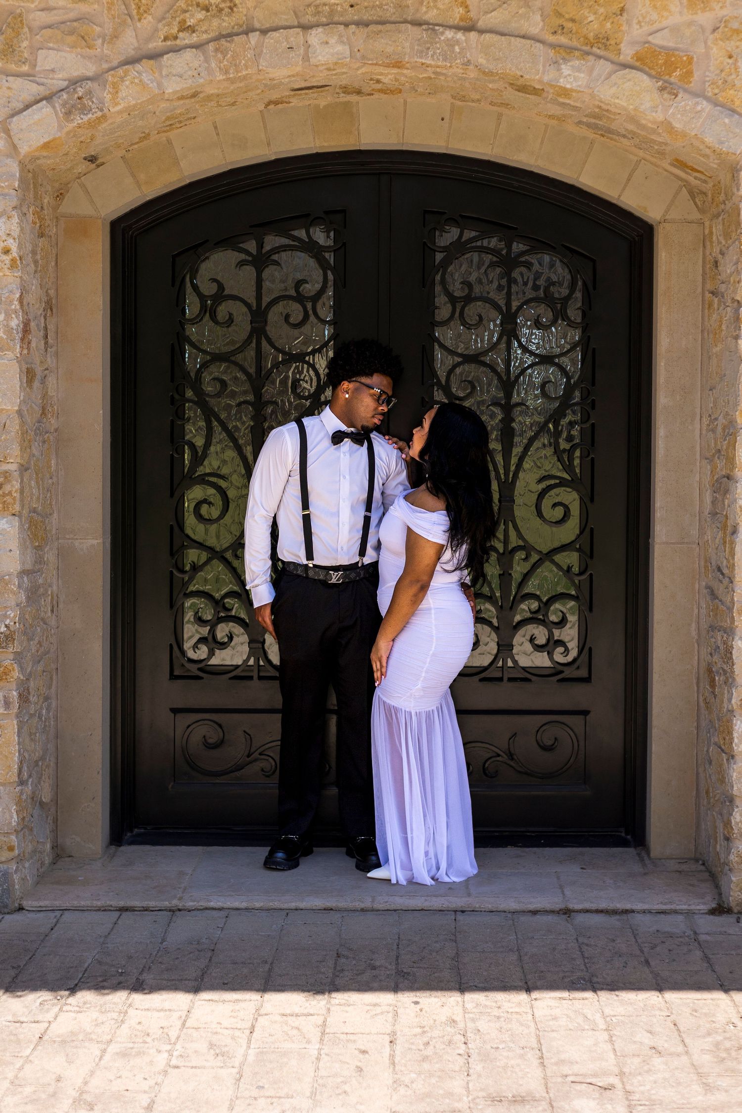 A couple in formal attire shares a romantic moment before ornate iron doors set in a stone archway.