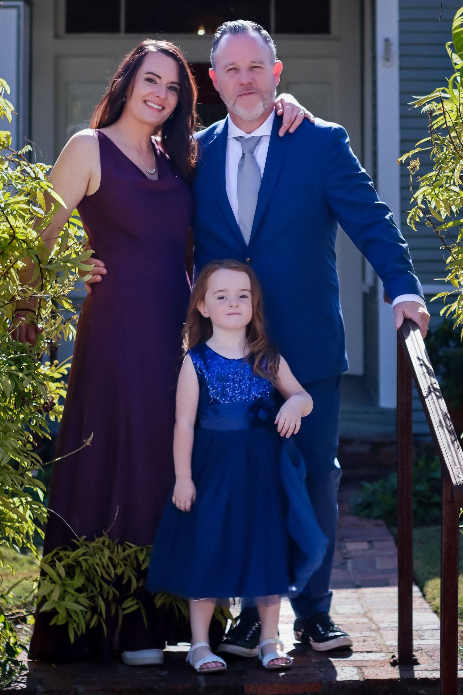 Family group stands on front porch steps wearing coordinated navy blue and purple formal outfits.
