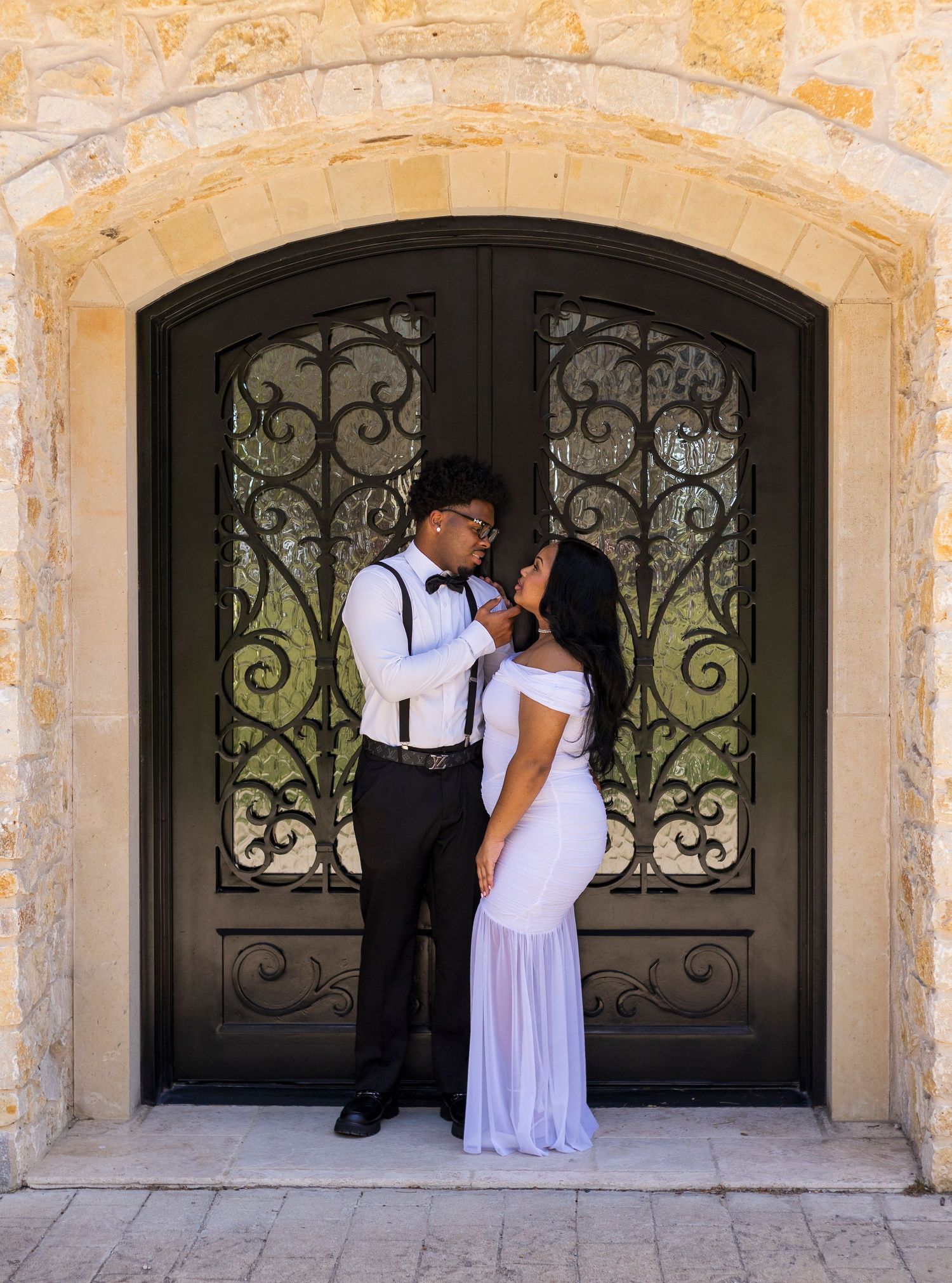 Couple posing in front of ornate black iron double doors framed by a stone arch, dressed in formal attire.