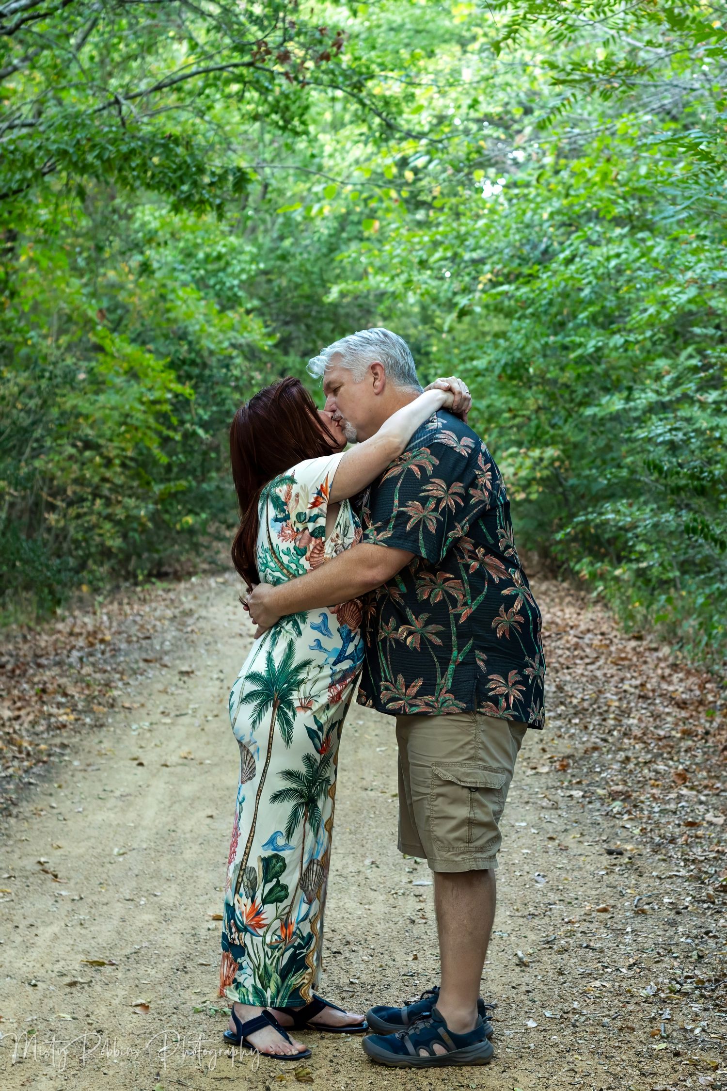 Middle-aged couple sharing a romantic kiss on a scenic forest path, wearing tropical print outfits.