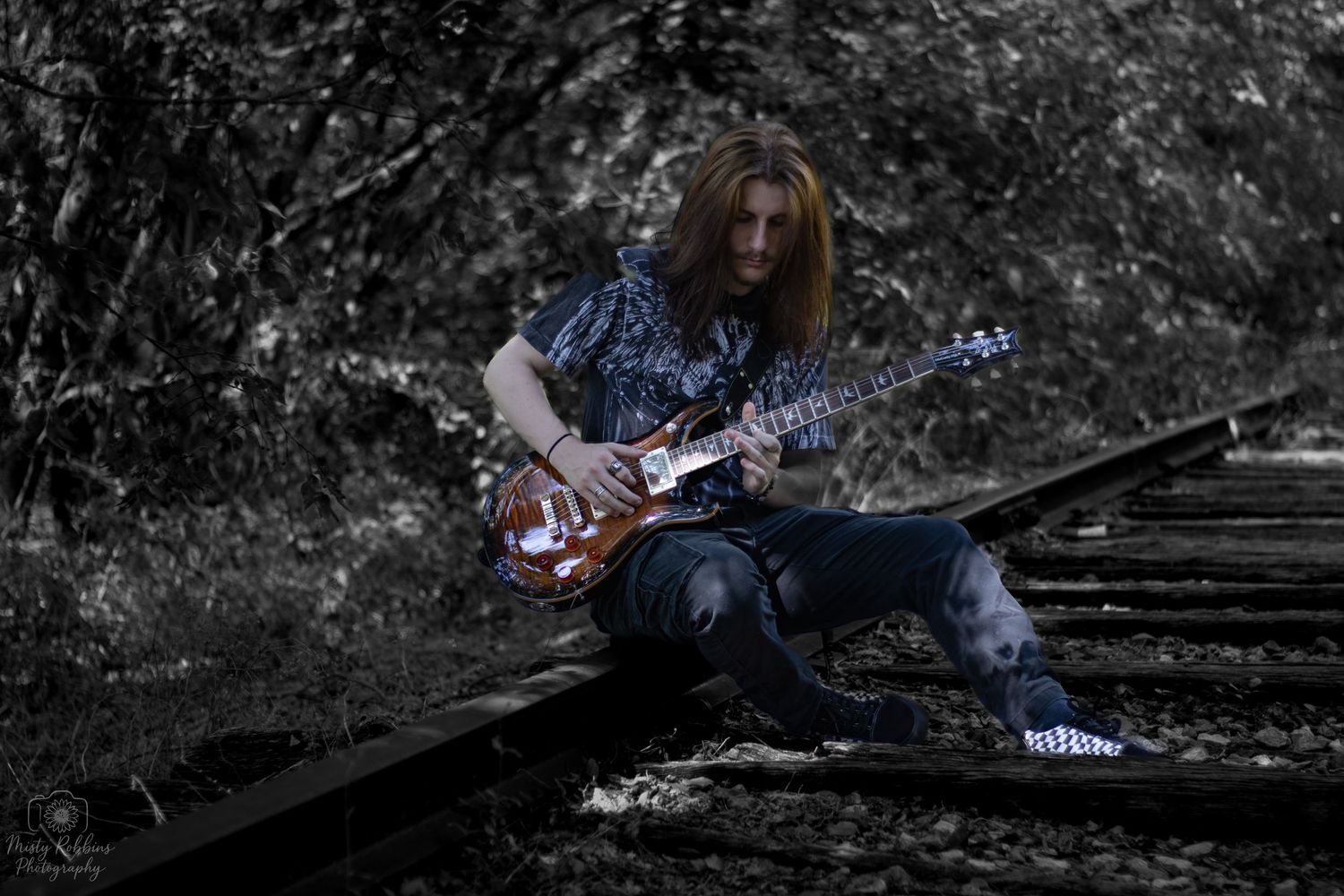Musician playing electric guitar while sitting on railroad tracks in a desaturated outdoor setting.