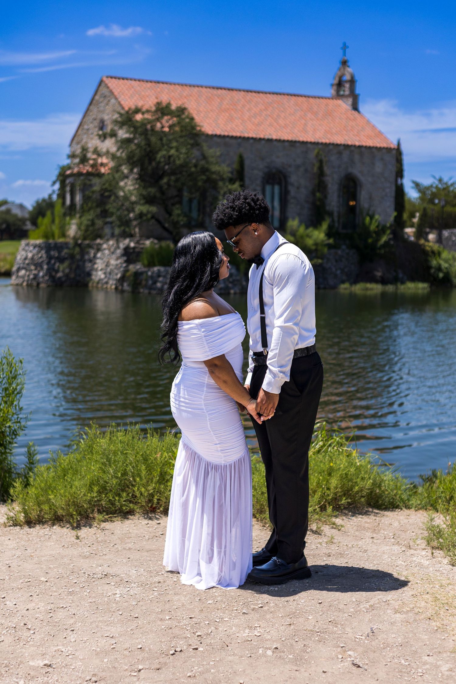 Couple in white dress and black suit holding hands by a lake with a stone chapel in the background on a sunny day.