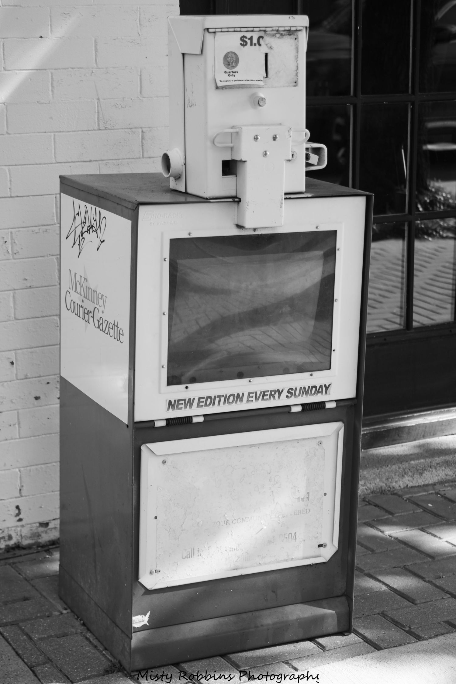 A row of vintage newspaper vending machines lined up against a white brick wall in black and white.