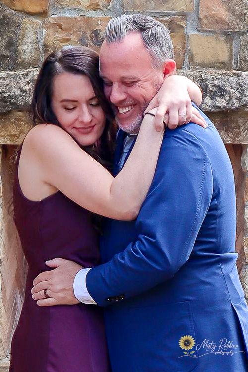 Couple embracing warmly in front of stone wall, woman in purple dress and man in blue suit smiling.