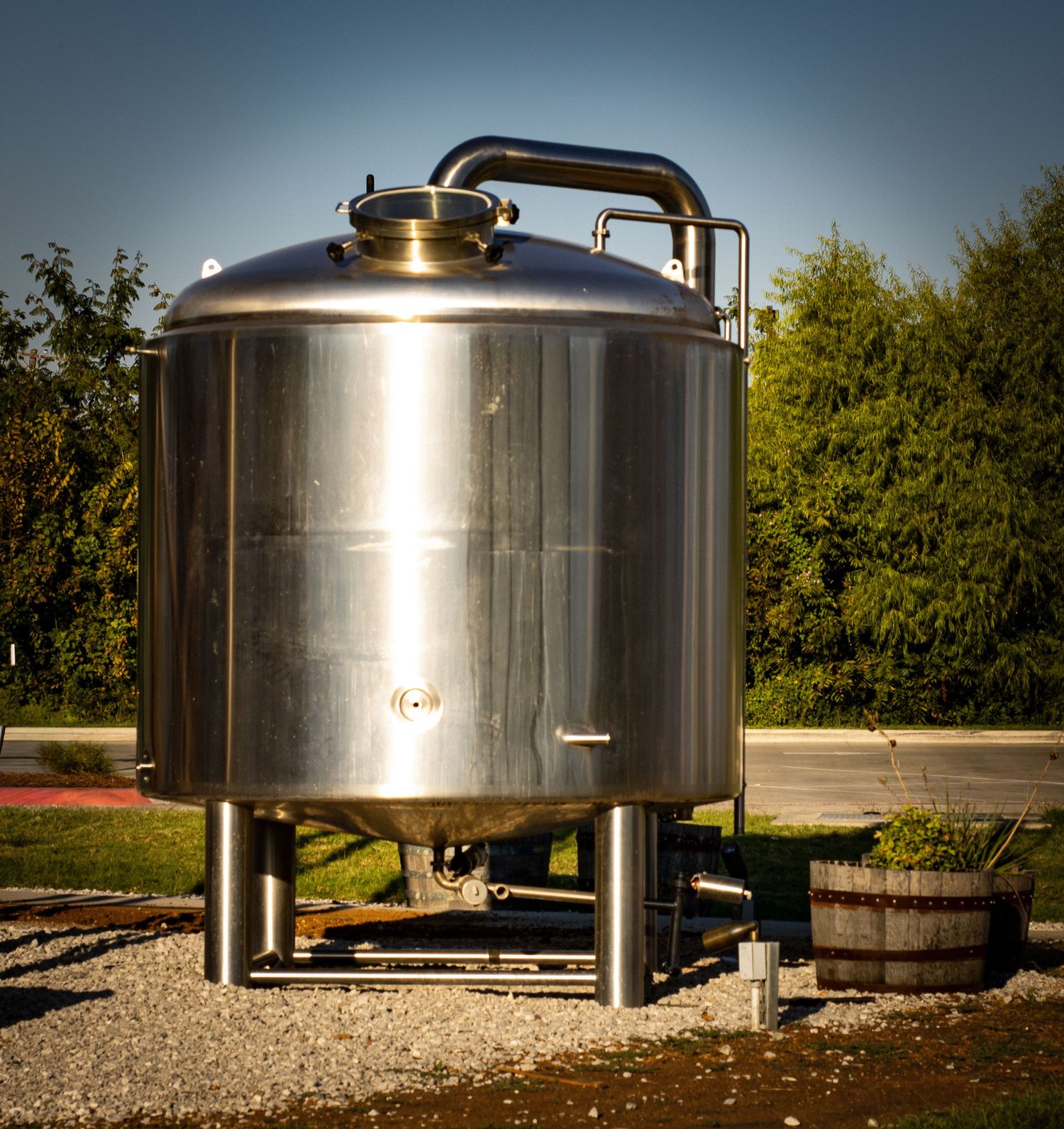 Row of stainless steel brewing tanks on gravel ground in outdoor setting.