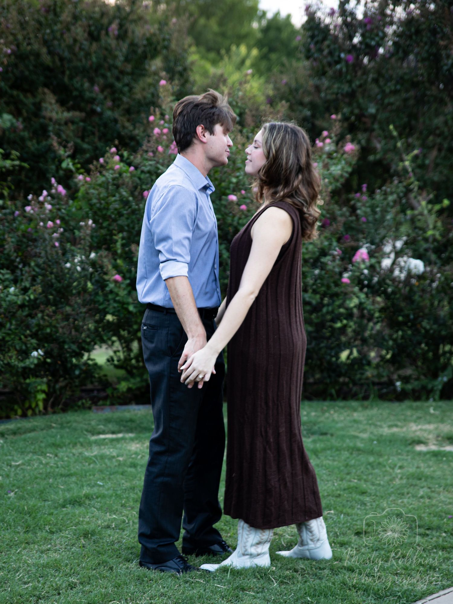A couple shares a tender moment while standing together on a grassy lawn surrounded by lush greenery.