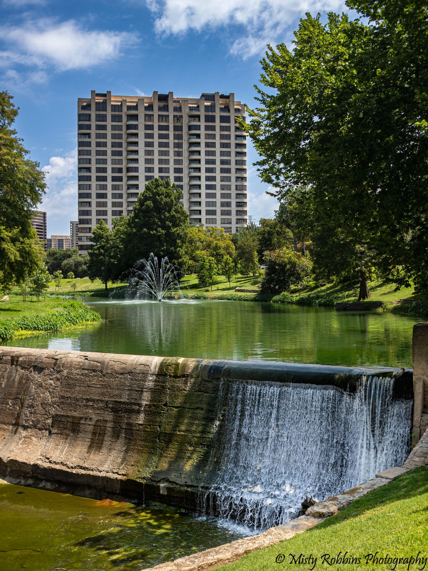 Urban park pond with fountain and waterfall cascade, lush green trees, and a tall modern building under blue skies.