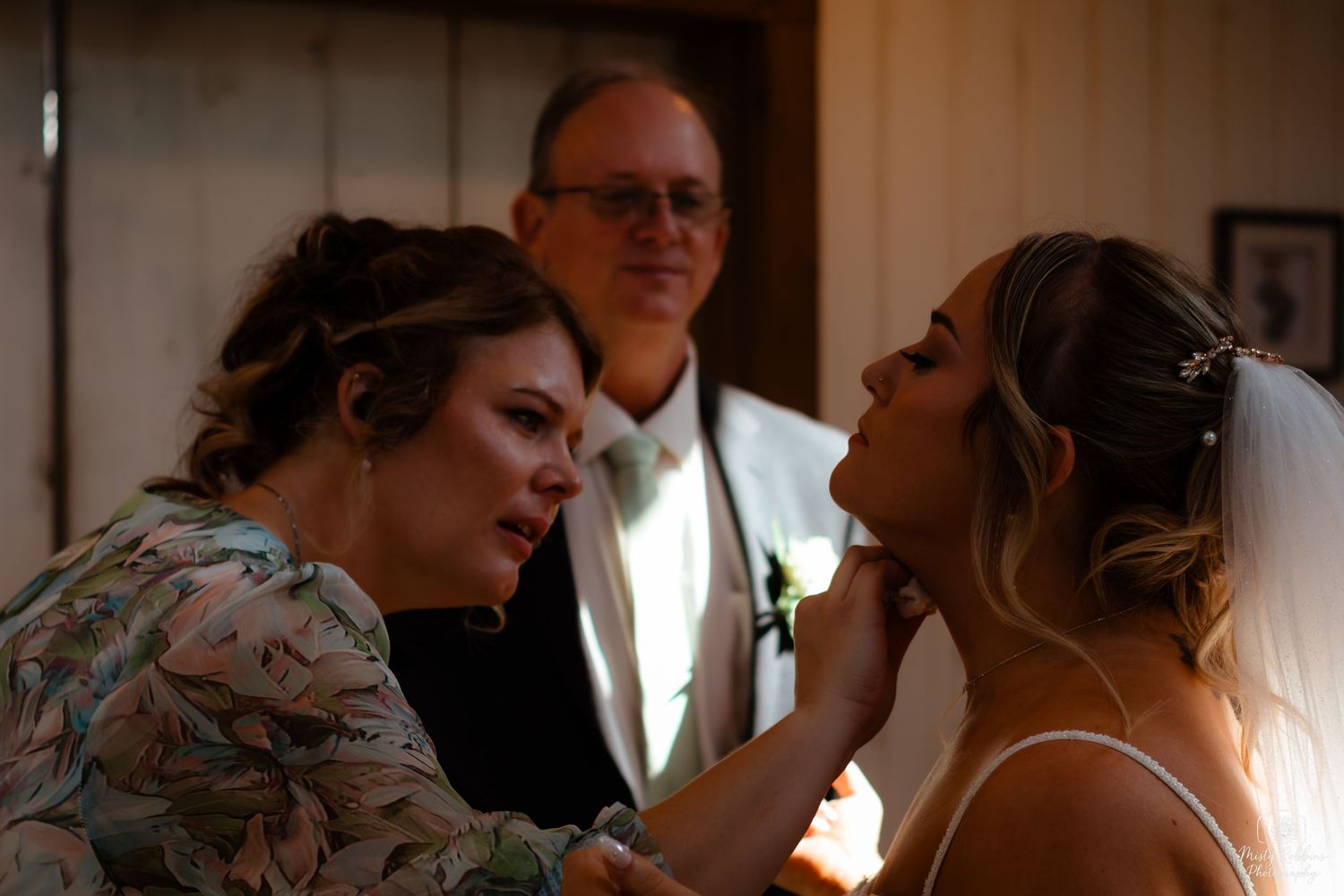 A woman helps a bride with makeup while a man in a suit watches in a warmly lit room before a wedding ceremony.