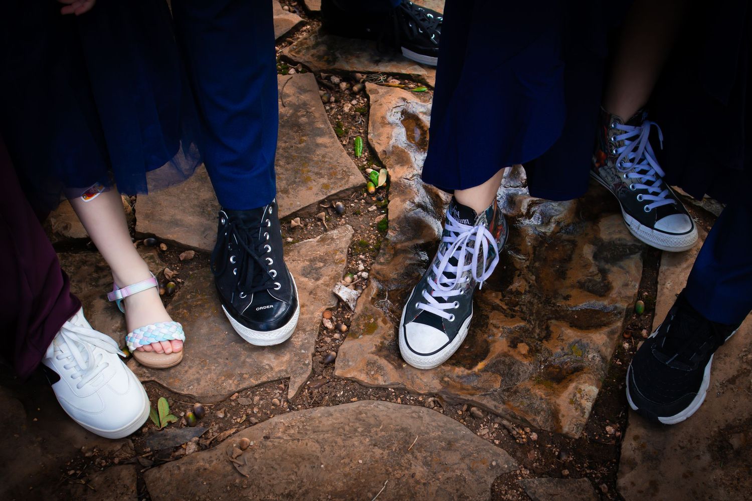 Multiple pairs of feet wearing sneakers and shoes standing on a rocky or cobblestone surface.