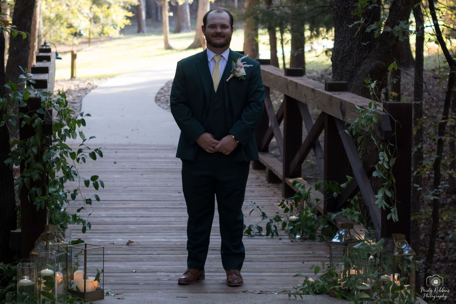 A person in formal attire stands on a wooden boardwalk surrounded by lush greenery and trees.