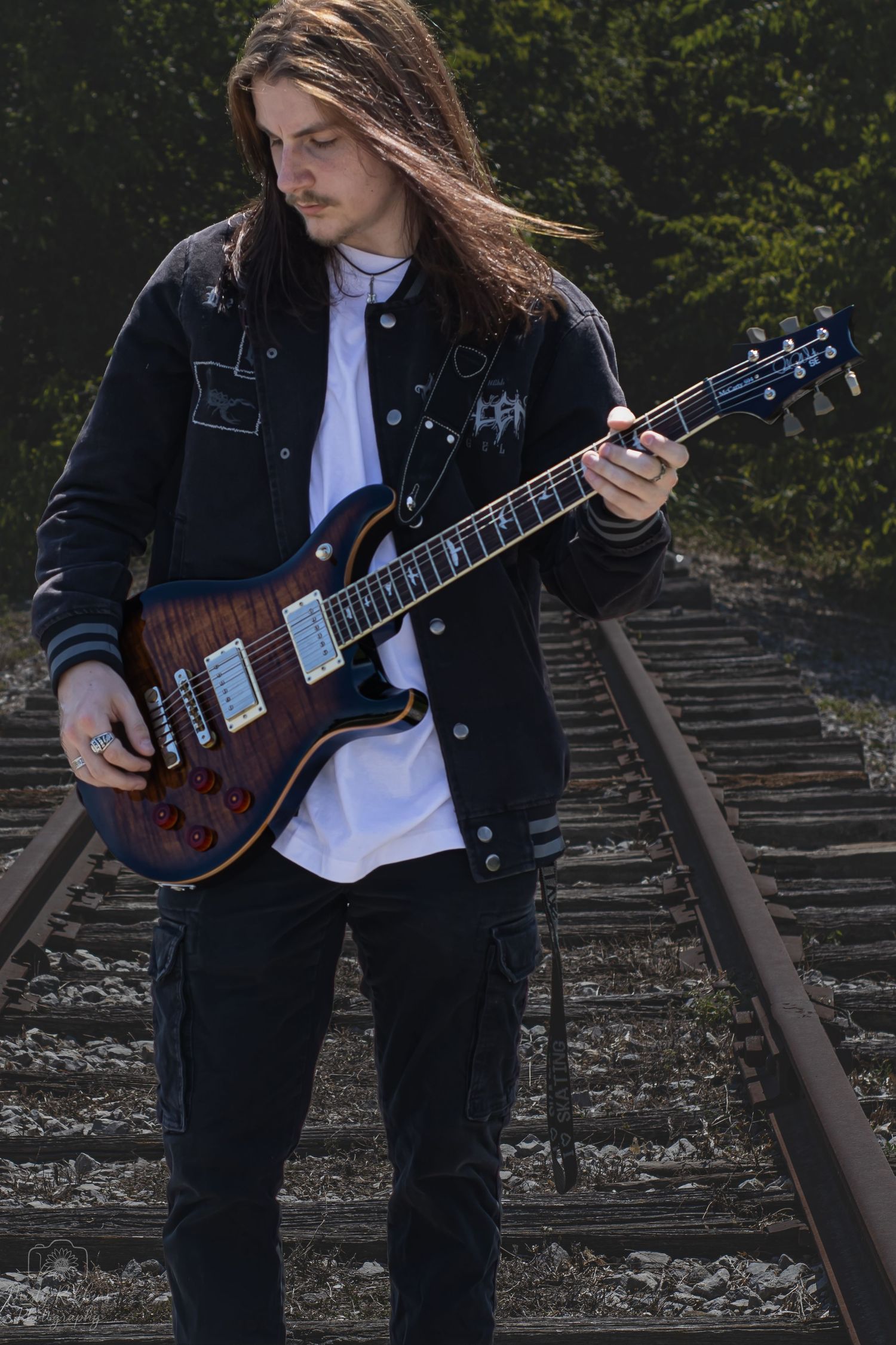 Musician in black jacket plays electric guitar while sitting on train tracks in artistic photo series.