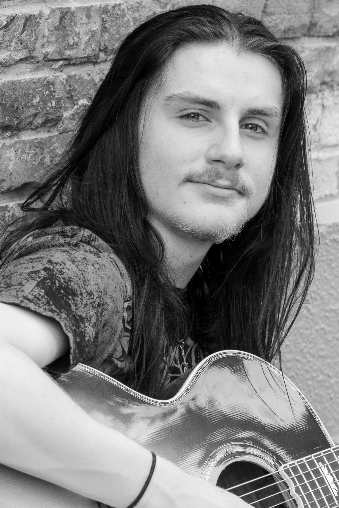 Young man with long hair playing acoustic guitar against brick wall, black and white portrait photo.