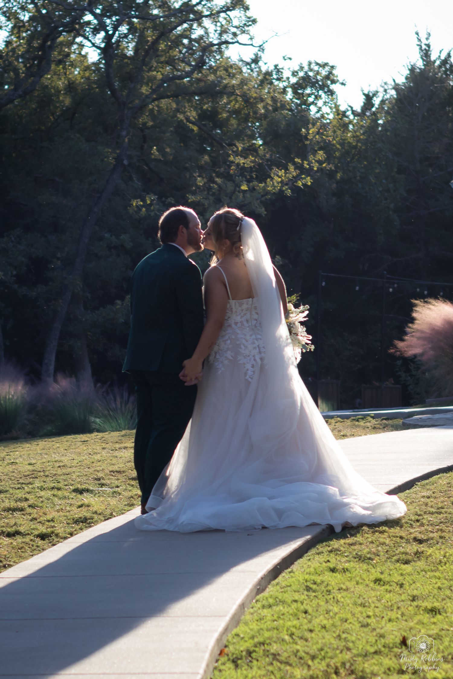 A couple in wedding attire sits together on a winding concrete path surrounded by green grass and trees at sunset.