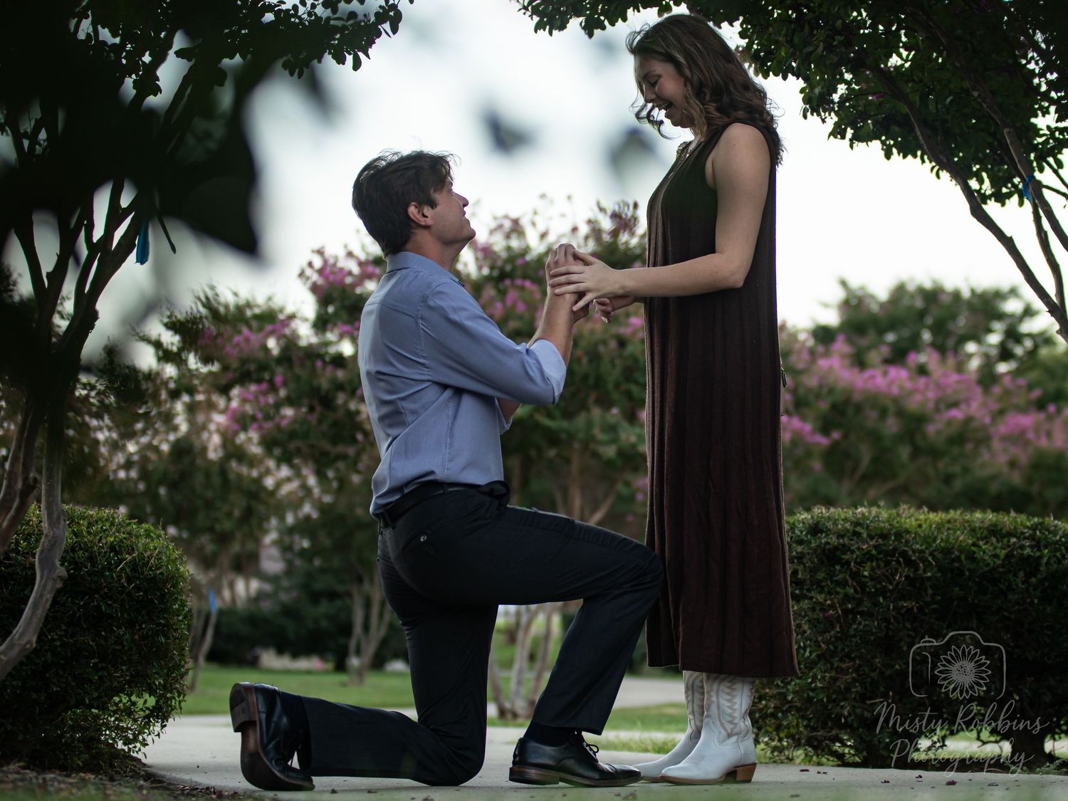 Man proposing to woman in a park, down on one knee holding a ring, surrounded by blooming trees.