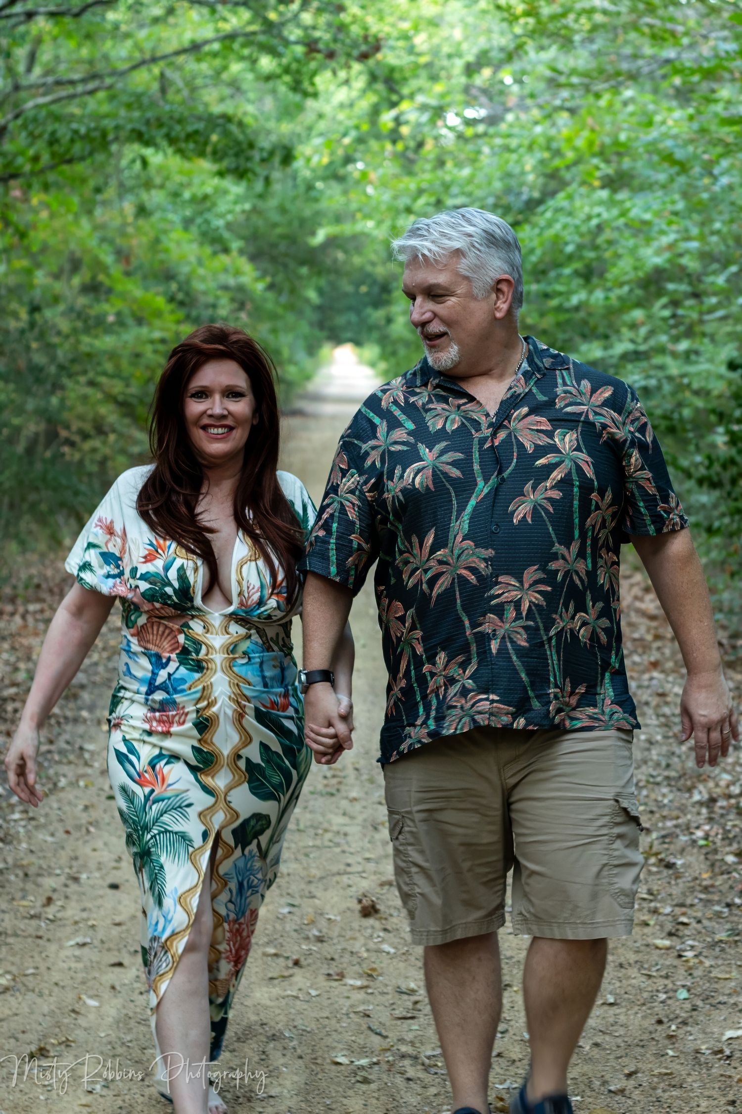 Couple holding hands walking on a tree-lined path, woman in floral dress, man in tropical shirt.
