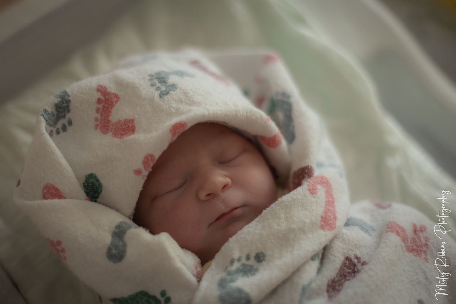 Newborn baby sleeping peacefully wrapped in a colorful footprint swaddle blanket with a hood in a hospital bassinet.