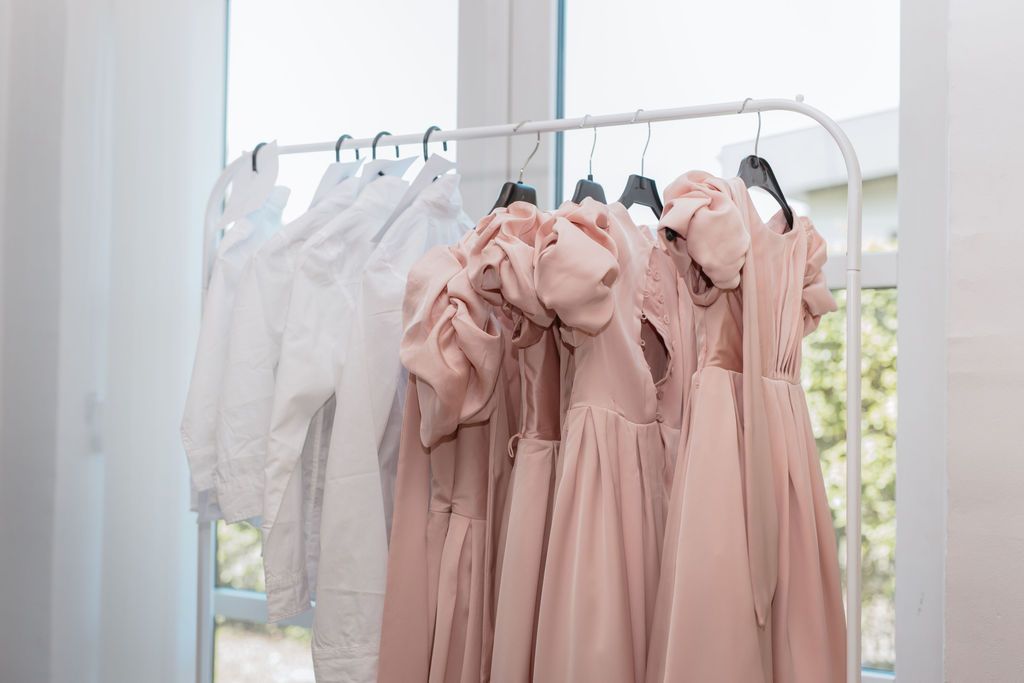 Blush pink bridesmaid dresses hanging in a row on a white clothing rack near a window.