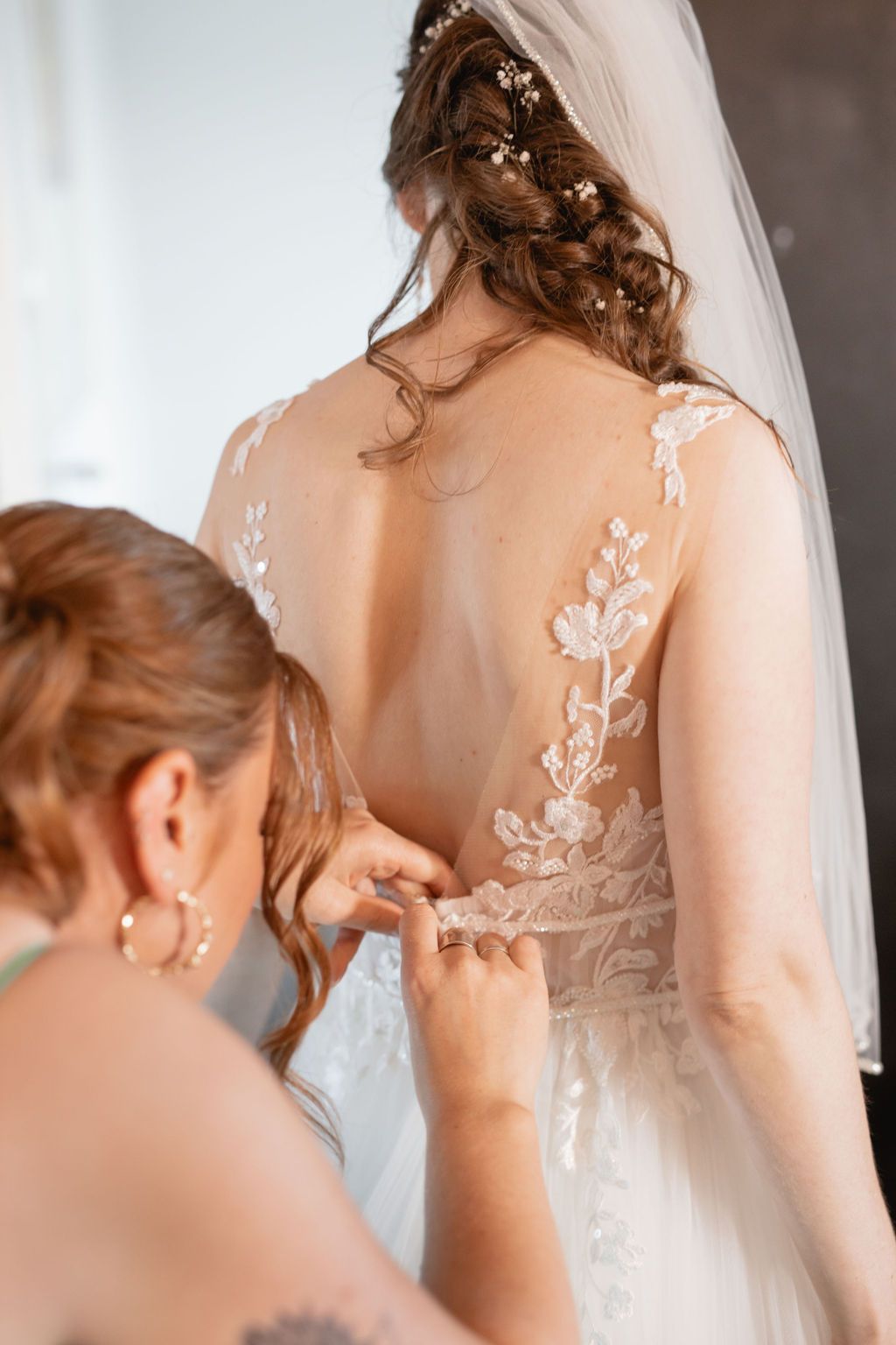 Back view of a wedding dress being buttoned up, showing intricate lace details and veil.