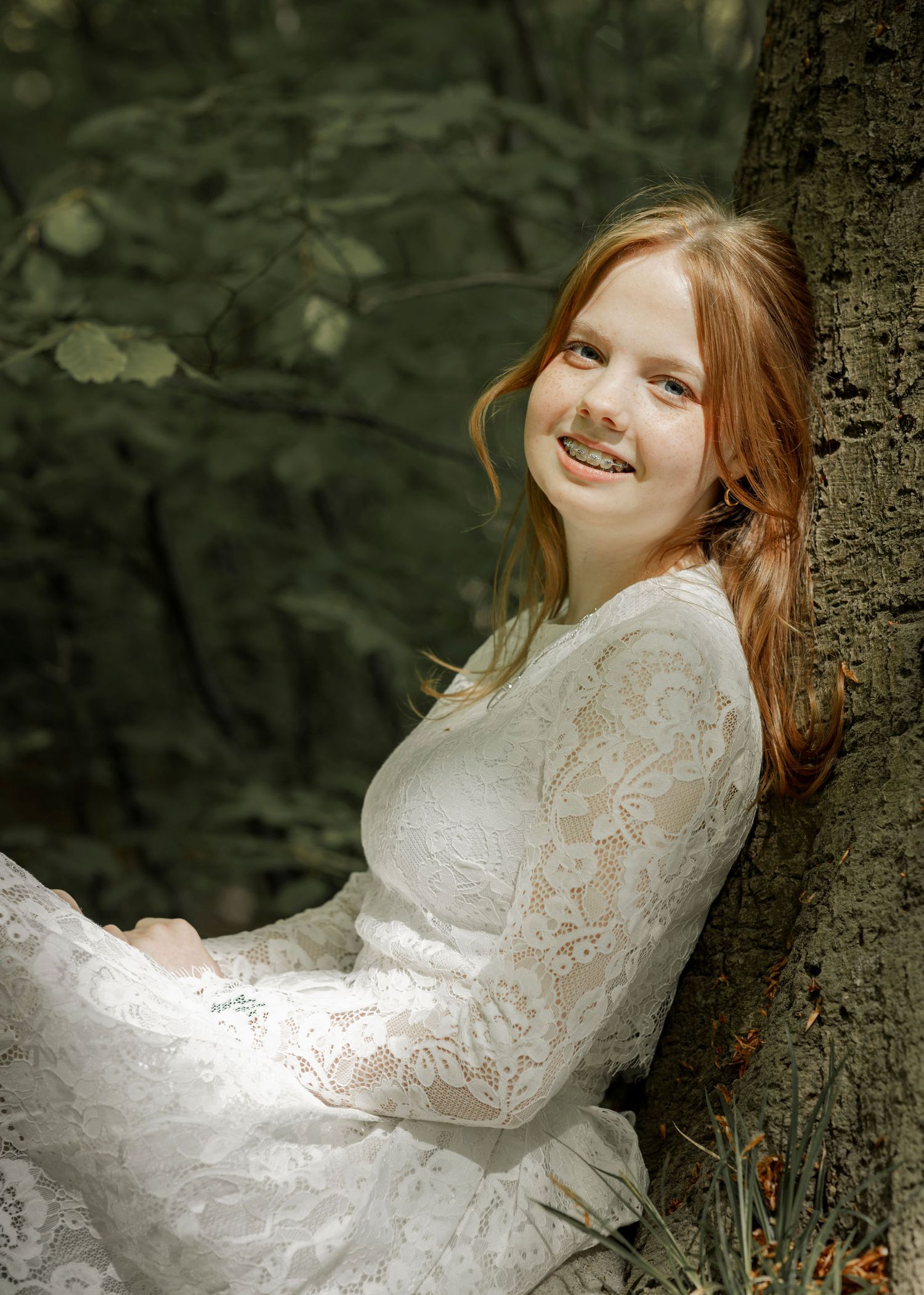 Person in white lace dress poses against tree in dark forest setting.