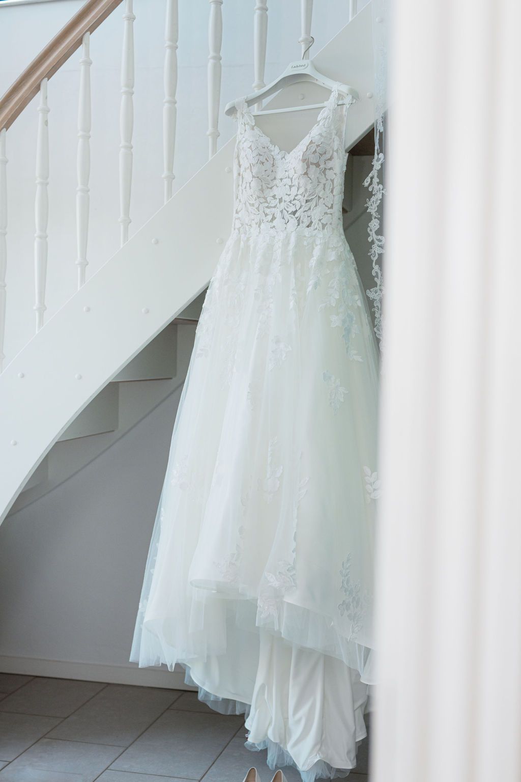 A delicate white wedding dress hangs elegantly against a white wall with stairs in the background.