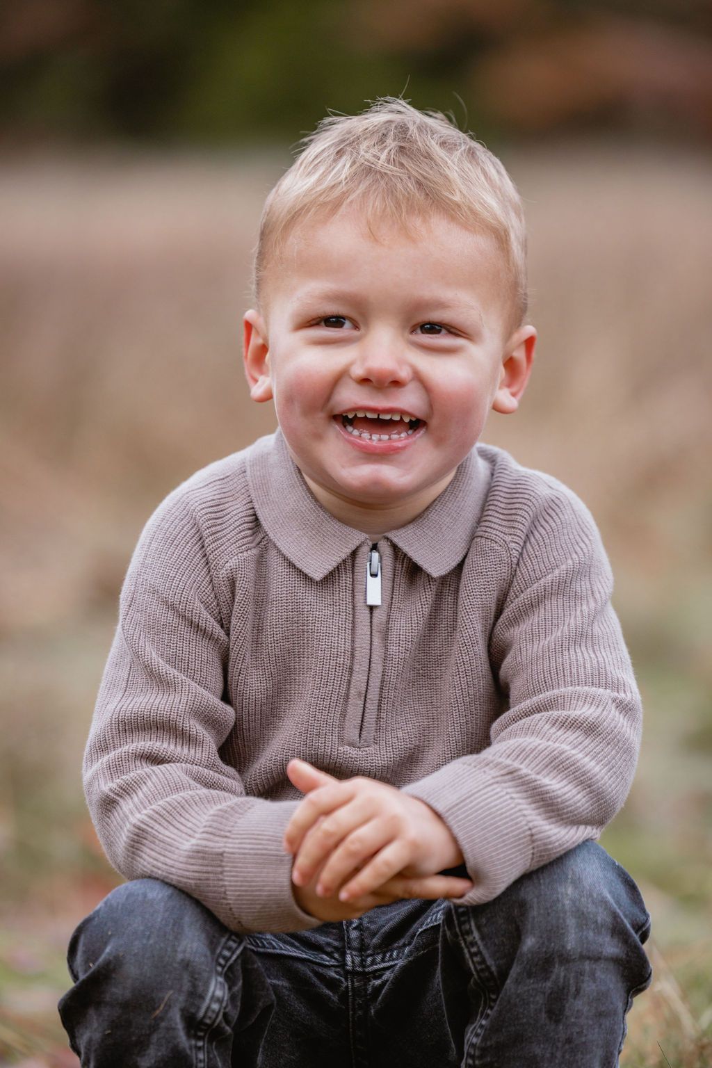 Child in grey sweater crouches and laughs with pure joy outdoors.