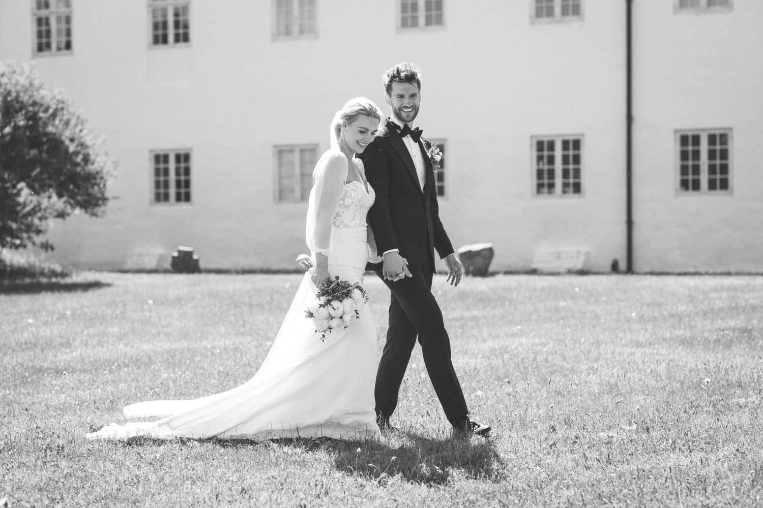 Black and white photo of newlyweds walking together on lawn near historic building.