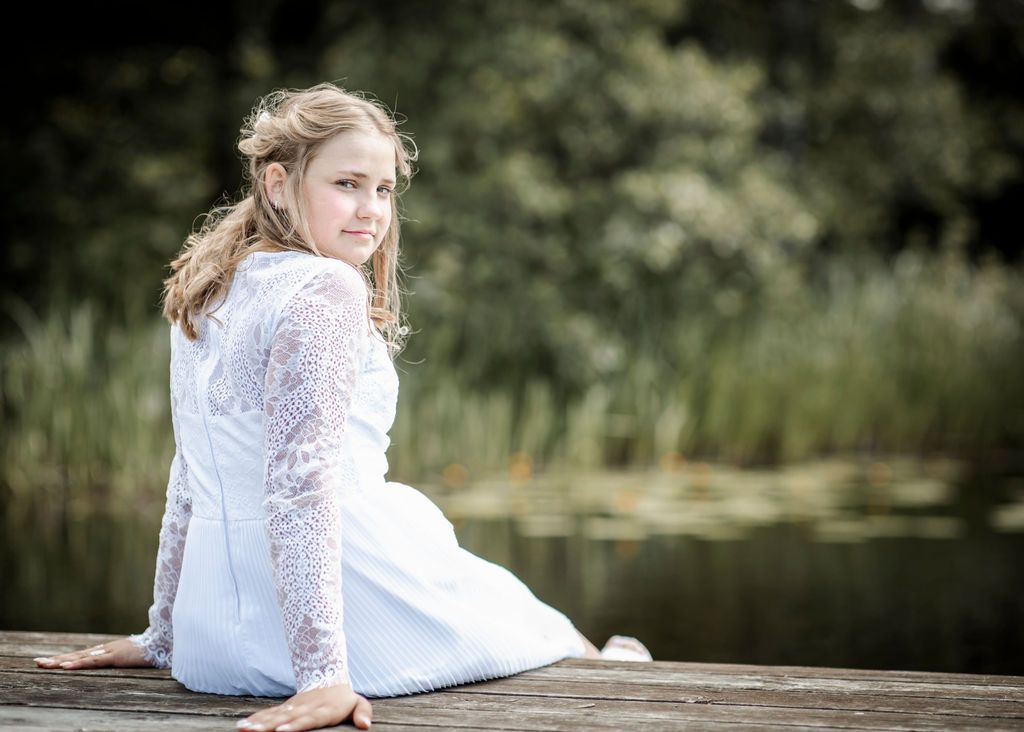 Person in white lace dress sits on wooden dock by lake during outdoor photo session.