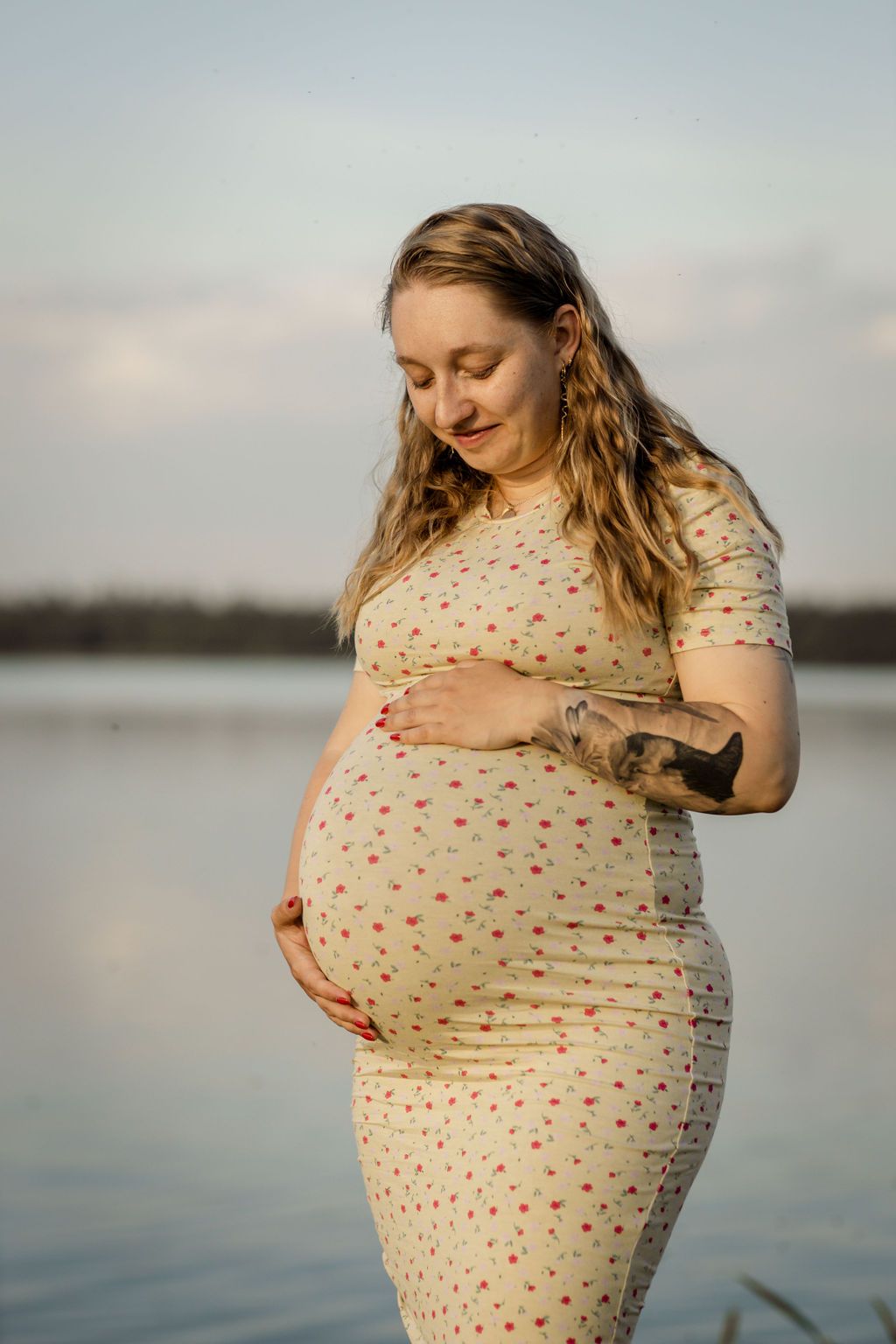 Expectant mother in polka dot dress poses by calm waters at sunset.
