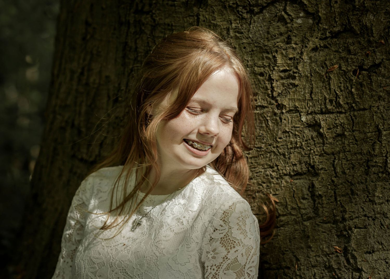 Person in white lace dress leans against textured tree bark in dramatic forest portrait.