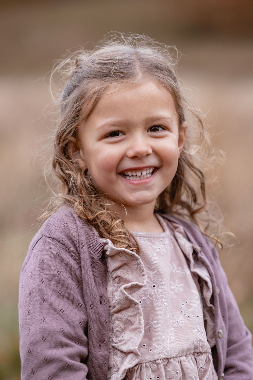 Young child with curly hair wearing purple cardigan smiles brightly at camera.