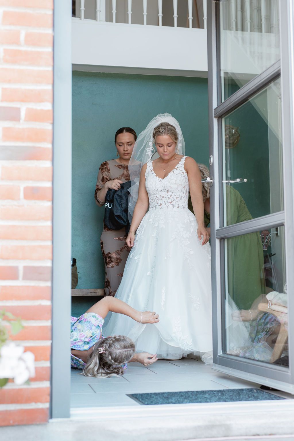 A bride in a white lace dress sits by a window in natural light.