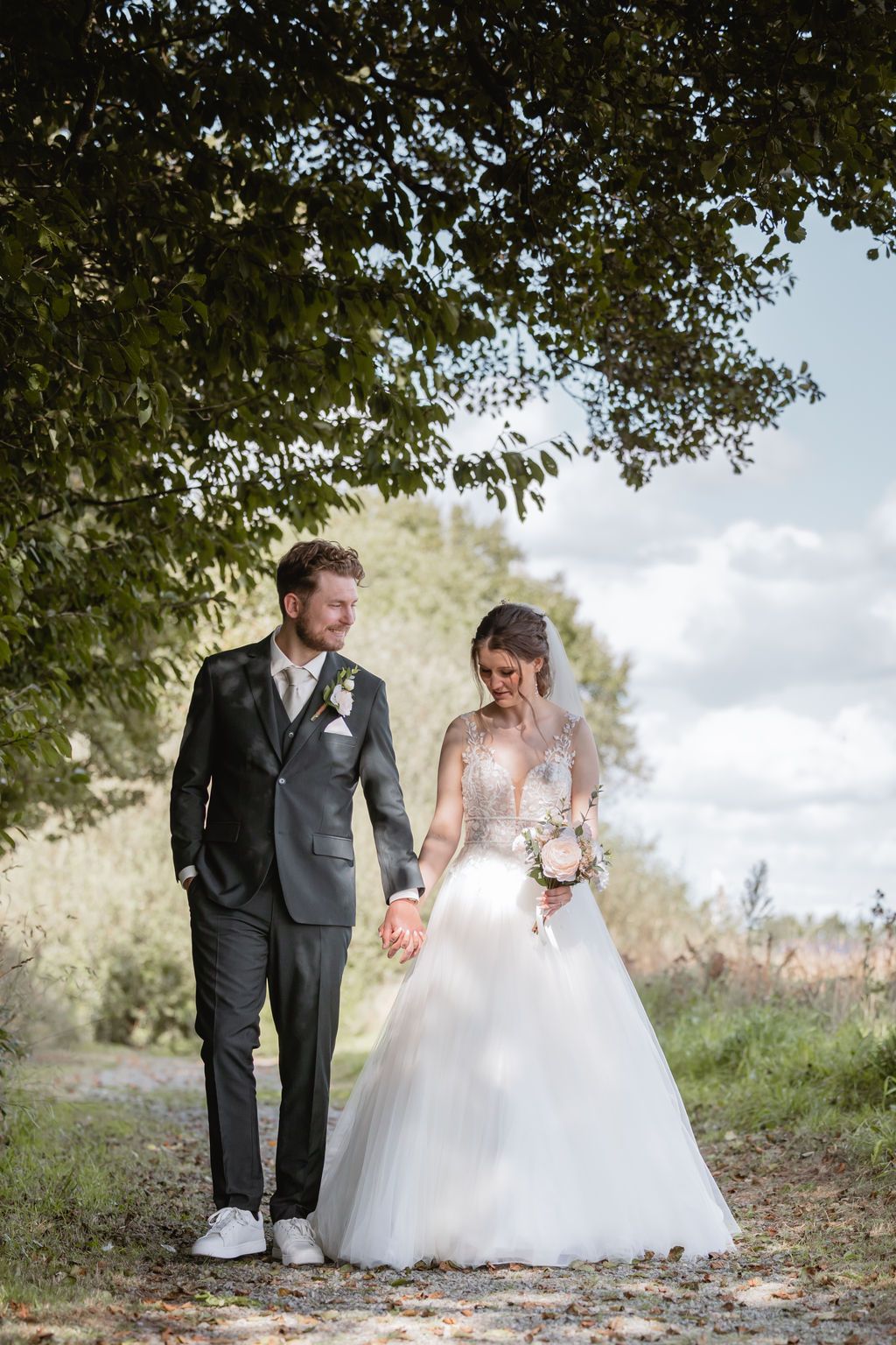 Newlyweds walking together on a tree-lined path in formal wedding attire.