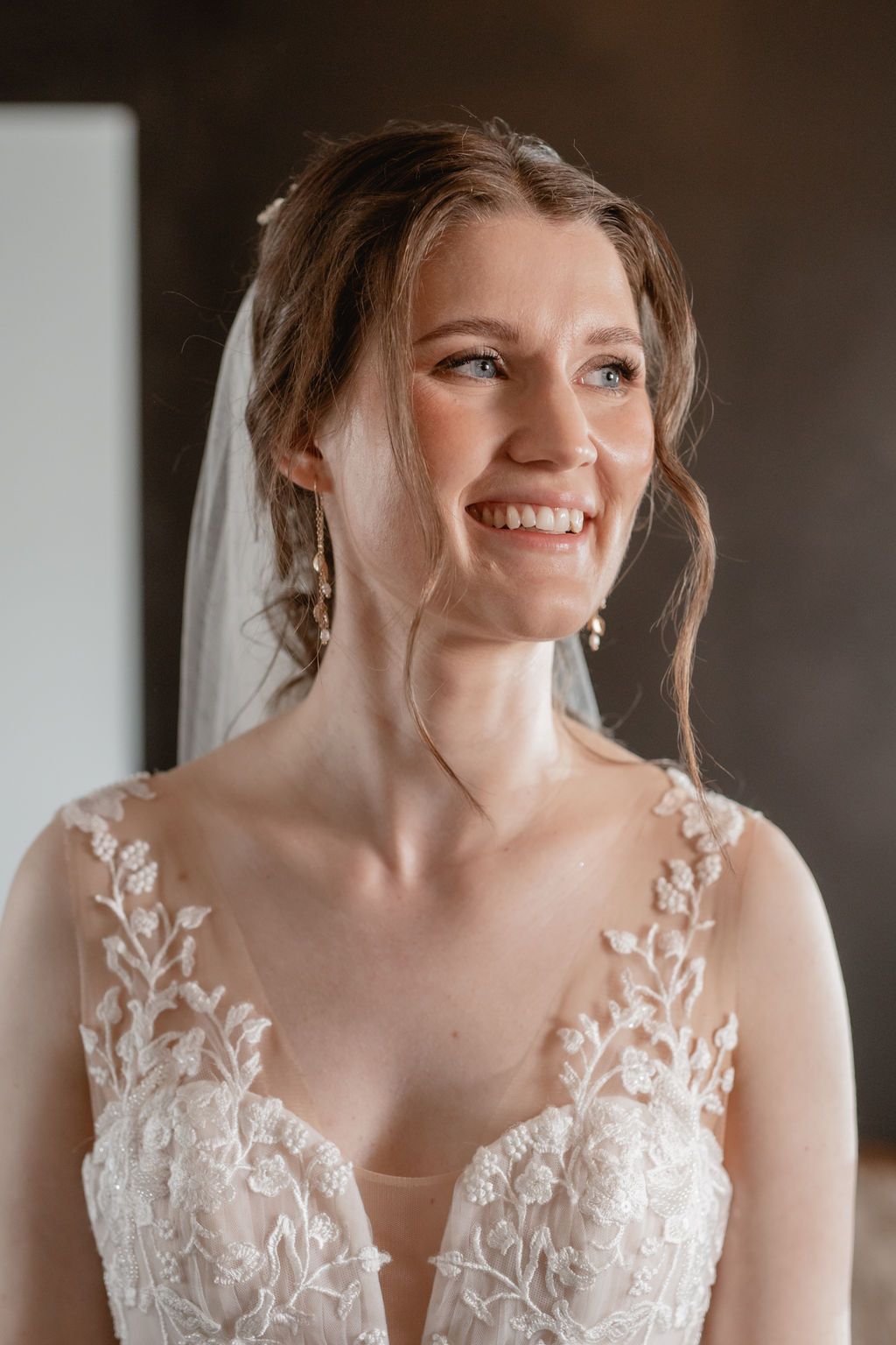 Bride wearing delicate lace wedding dress with floral appliques and sheer neckline smiles in natural light.