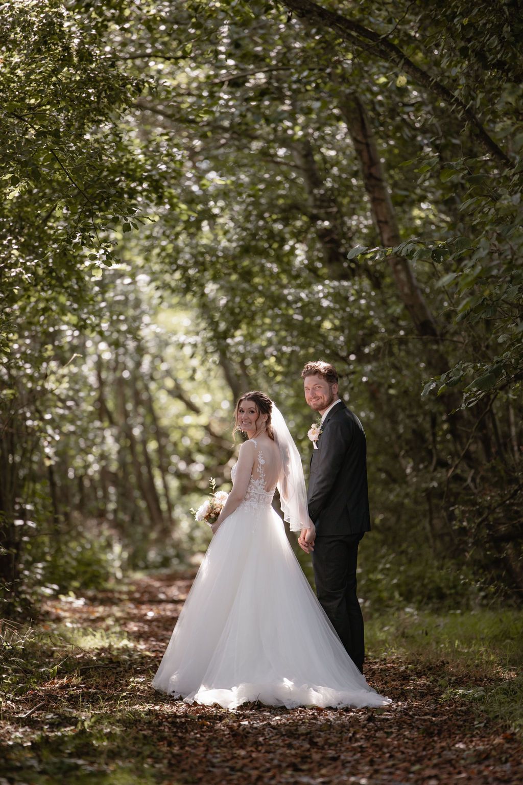Wedding couple poses in a romantic wooded setting with natural light filtering through trees.