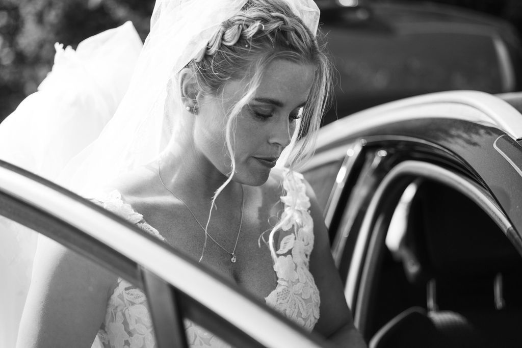 Black and white photo of a bride in a lace dress entering a car after her wedding.