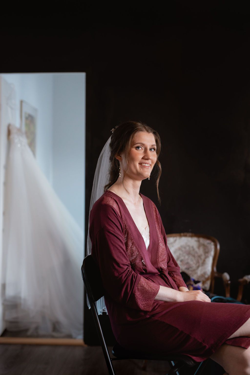 A person in a burgundy cardigan sits in preparation for a wedding with a dress visible in background.