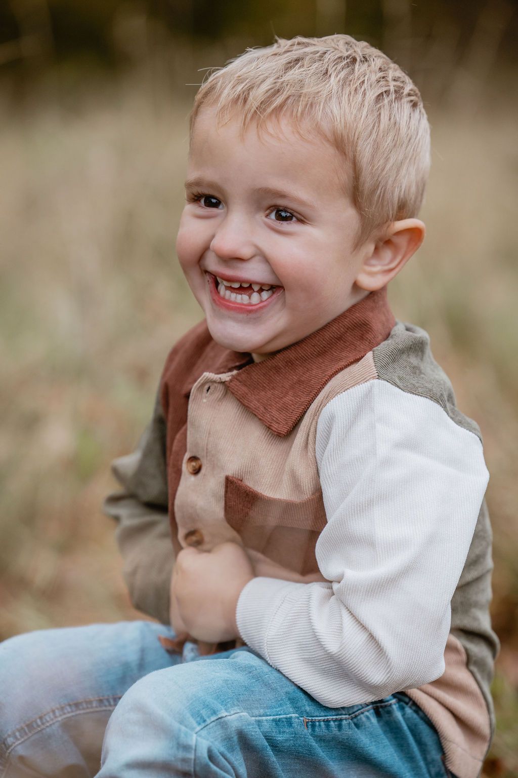 Young child wearing a brown vest and white shirt smiles brightly while sitting outdoors.