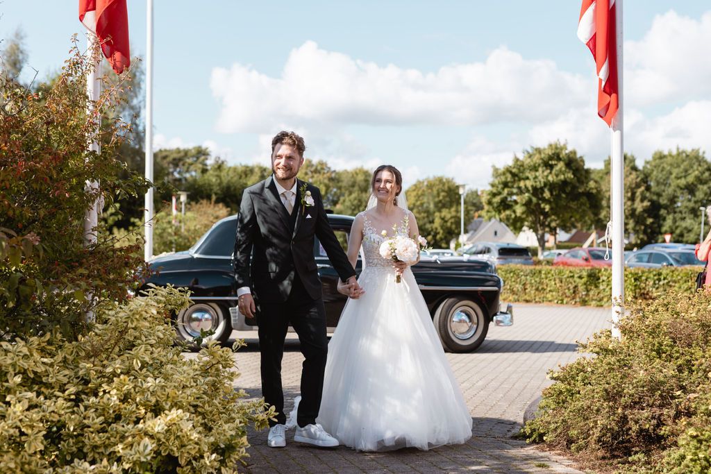 A couple walks together on a path with red flags and a classic car in the background.