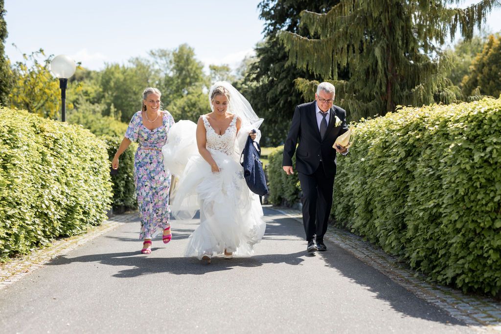 Wedding party walks down a garden path between manicured hedges on a sunny day.