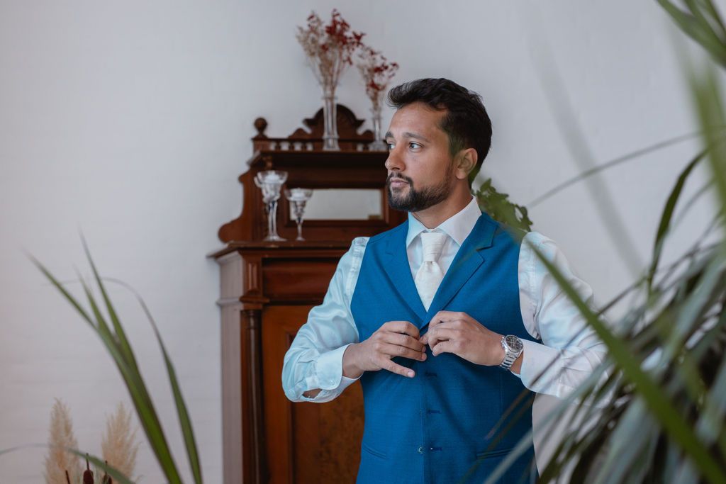 A person in a blue vest adjusts their attire while getting ready near an antique wooden cabinet.
