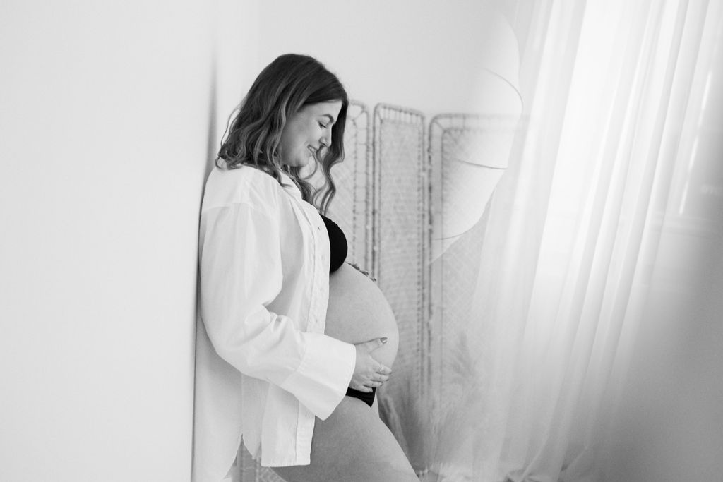 Black and white maternity portrait in front of sheer curtains.