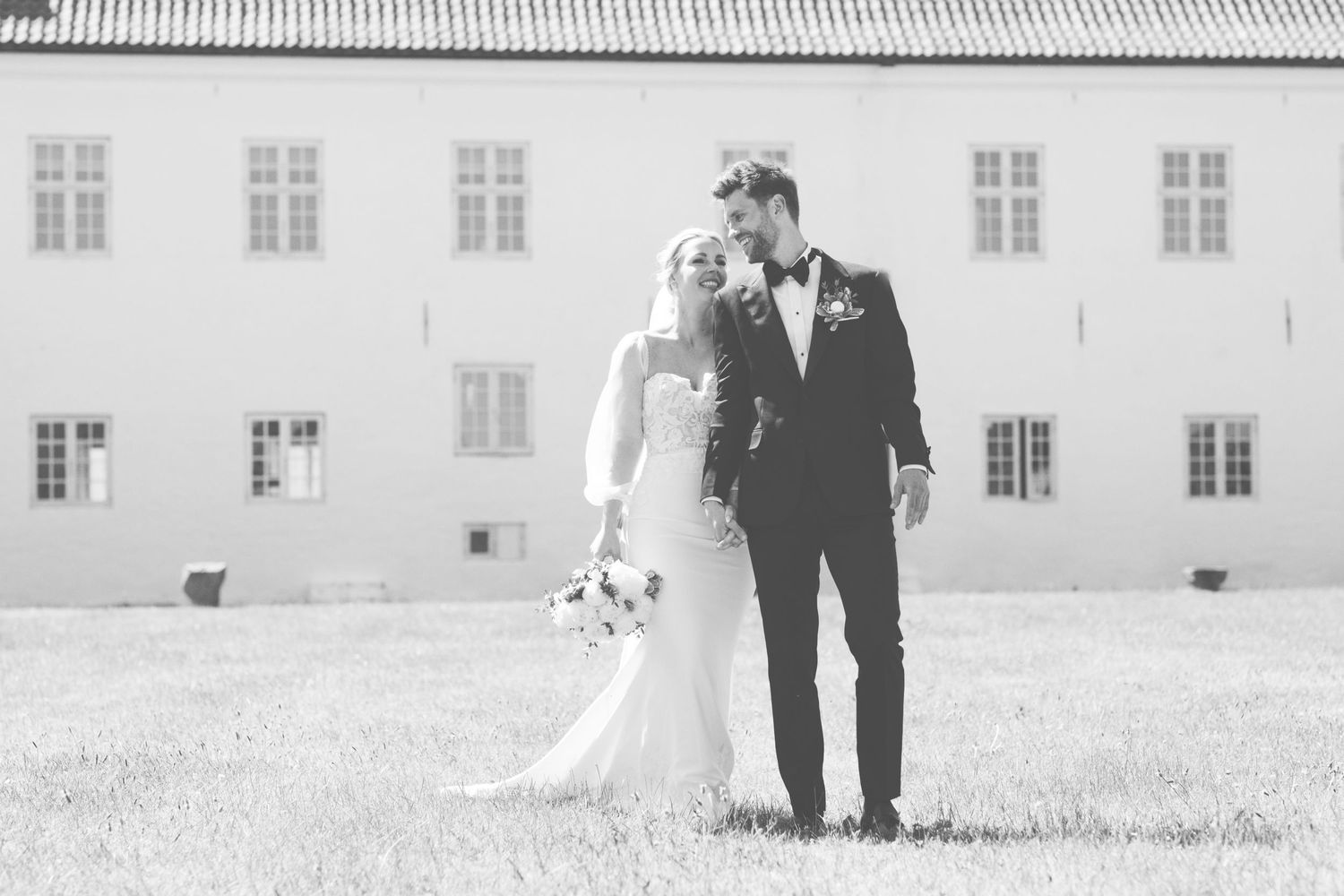 Wedding couple walking together on grass with white building in background.