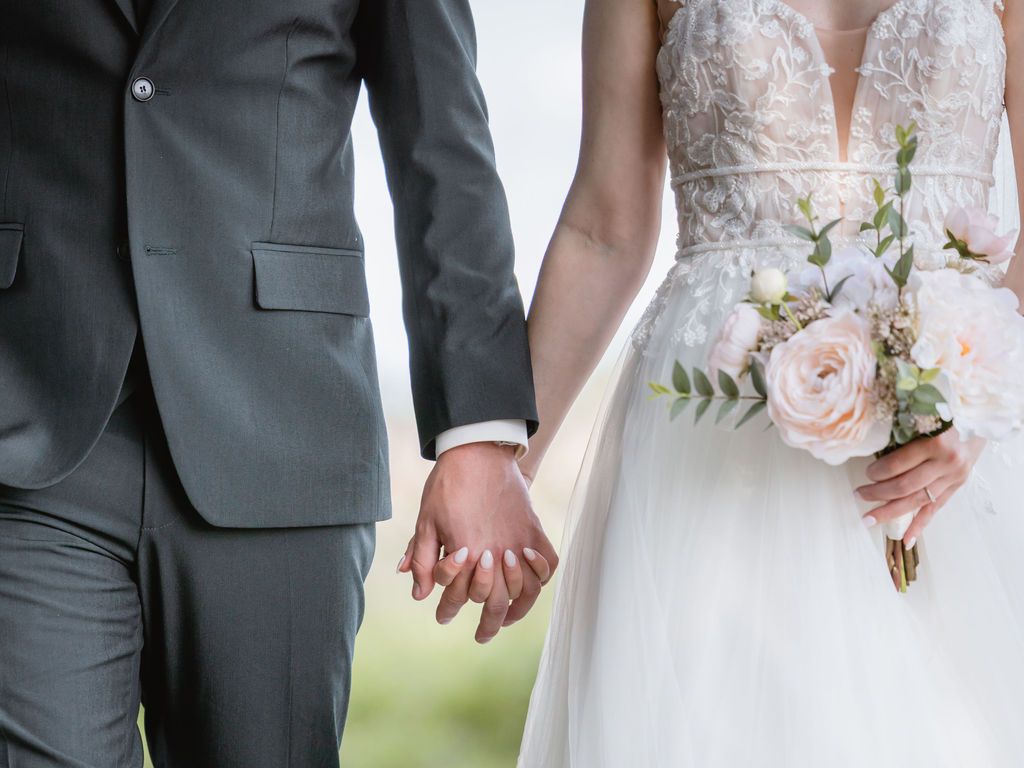 Closeup of bride and groom holding hands, showing wedding dress detail and gray suit.