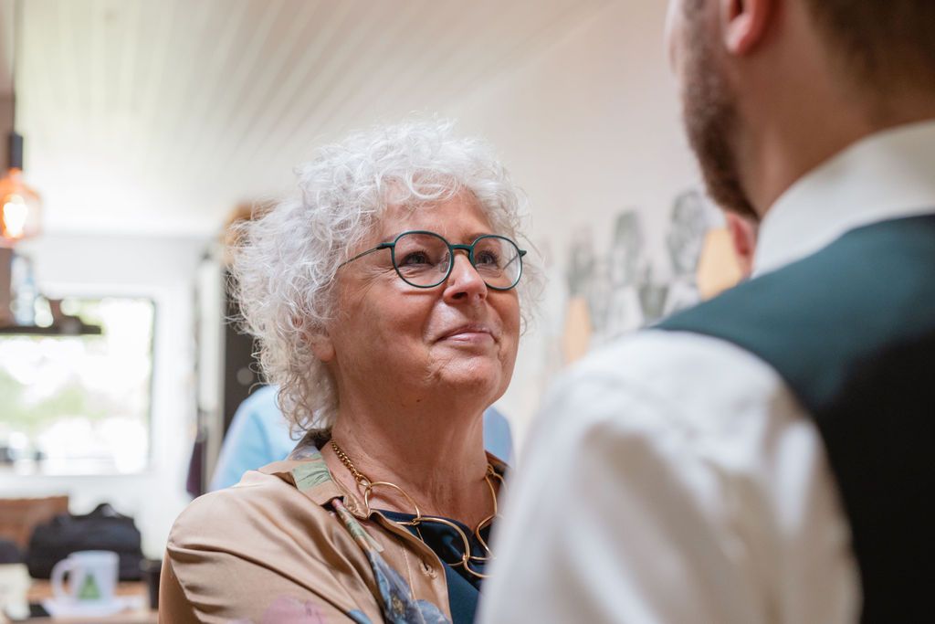 An elderly person with curly silver hair and glasses engages in conversation at an indoor event.