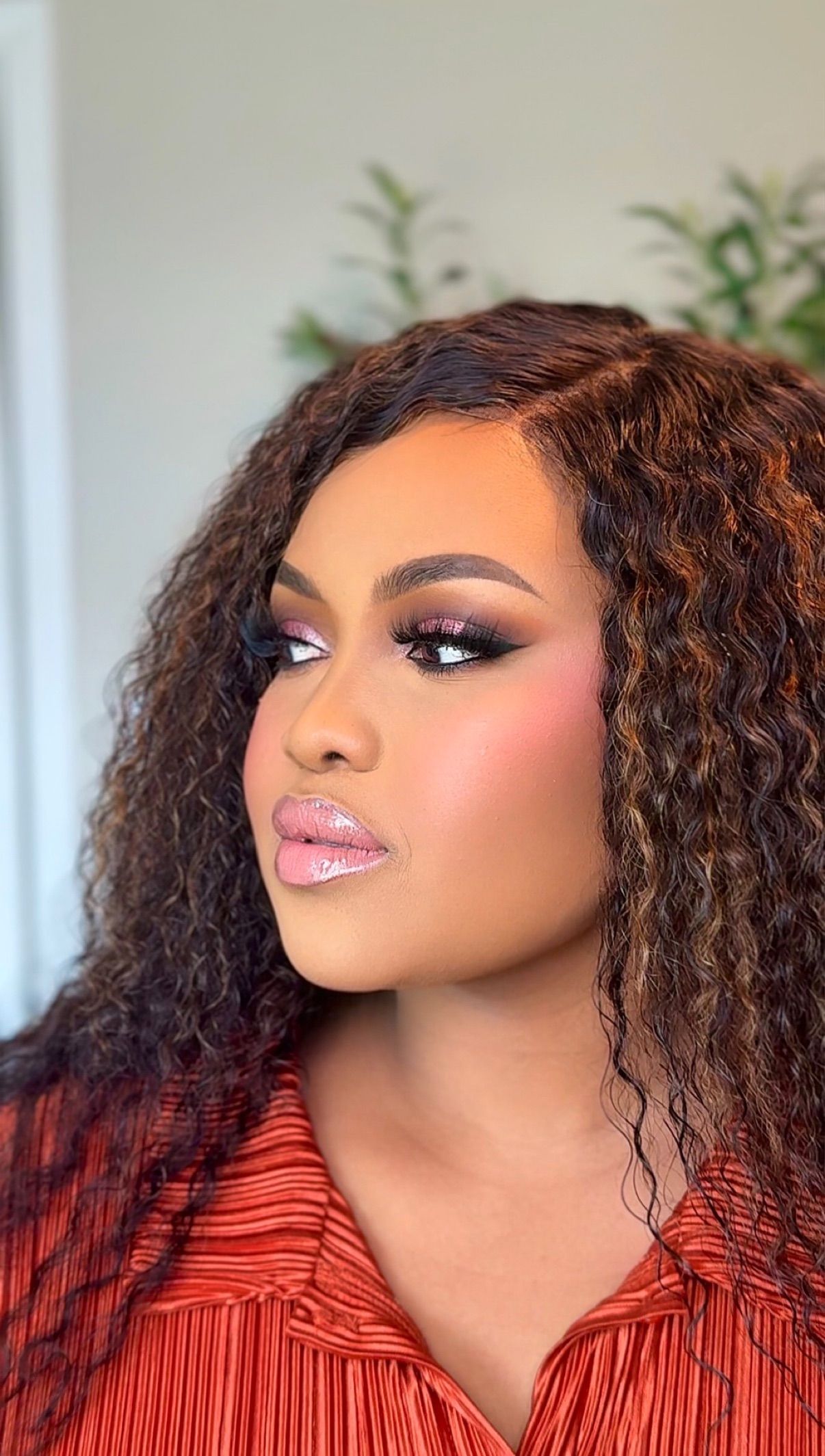 Woman with curly auburn hair wearing glamorous smoky eye makeup and glossy lips in a rust-colored top.