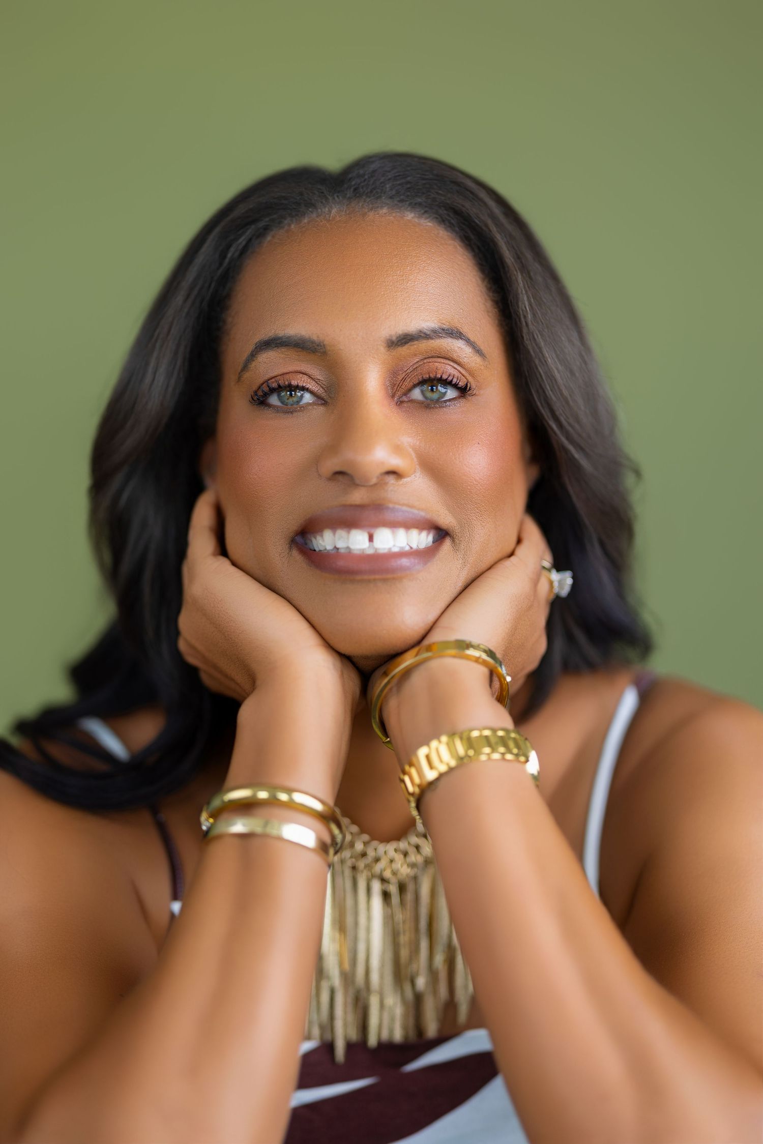 Smiling Black woman with gold bracelets and watch posing against olive green background.