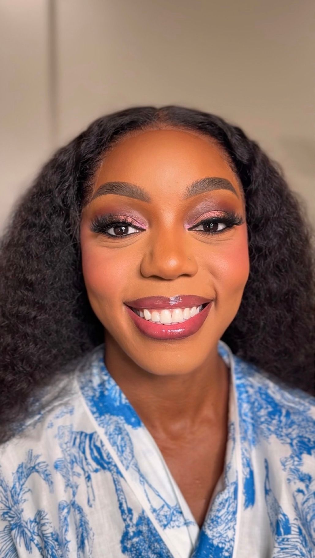 Smiling Black woman with glam makeup, pink eyeshadow, glossy lips, and curly hair wearing a blue floral top.