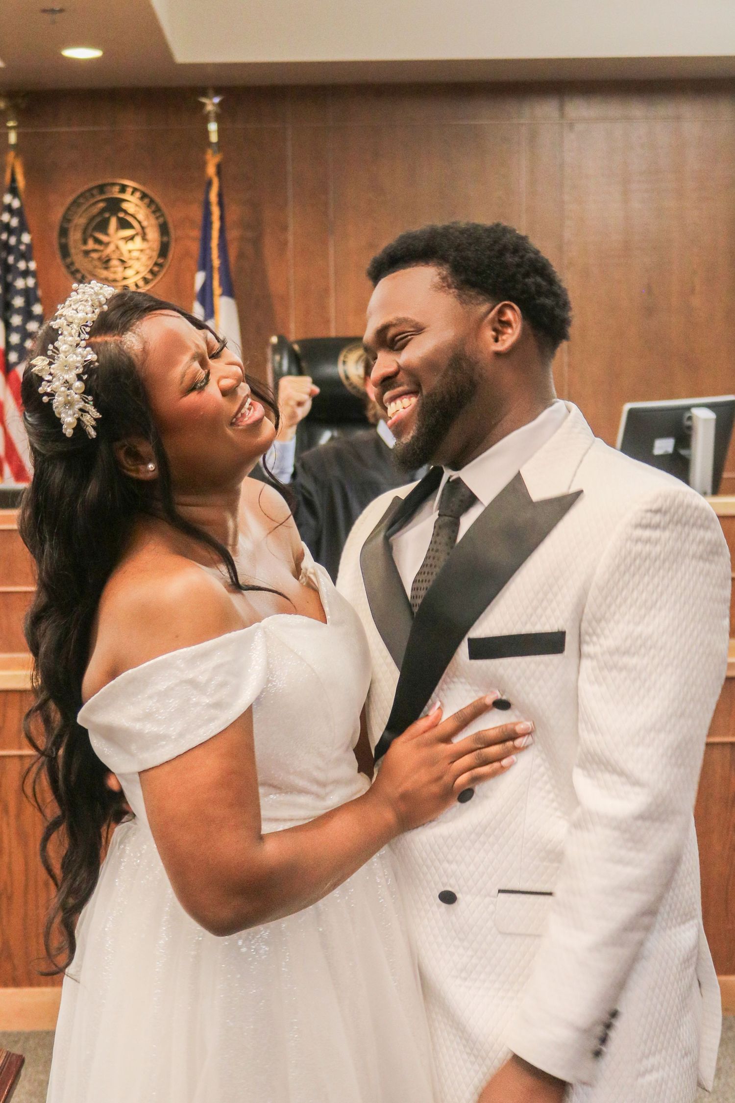 Joyful newlywed couple smiling at each other during courthouse wedding ceremony, bride in white gown, groom in white tuxedo.