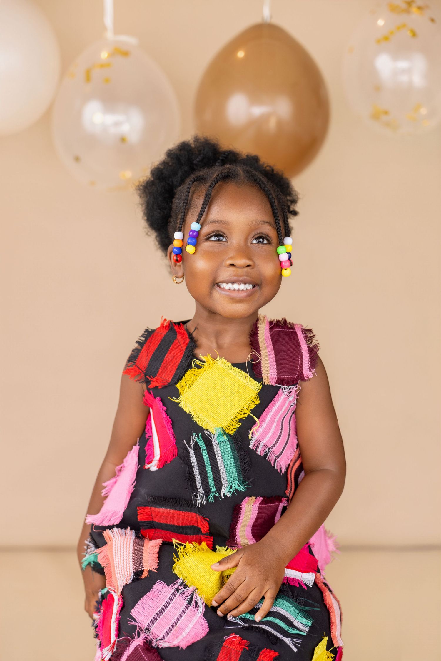 Smiling young girl in colorful patchwork dress with beaded braids, posing in front of balloons at a party.